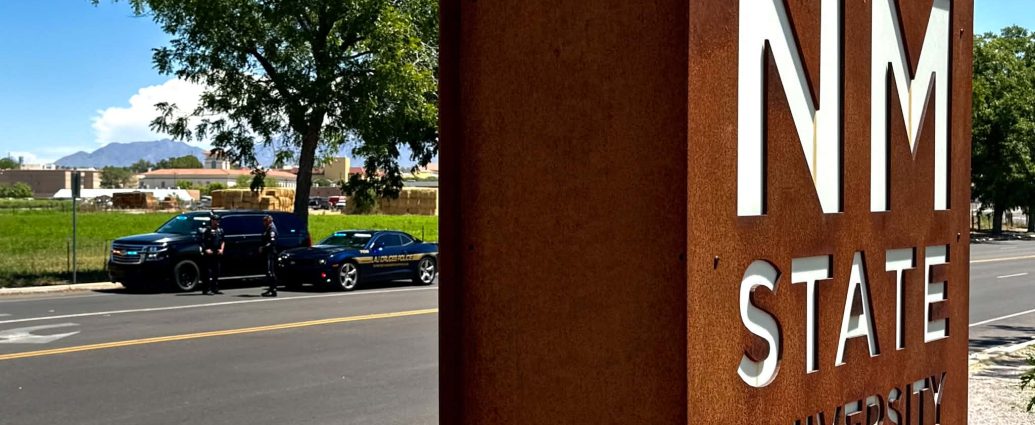 New Mexico State University sign near University Avenue, with Las Cruces Police and New Mexico State Police vehicles conducting a traffic safety operation in the background.