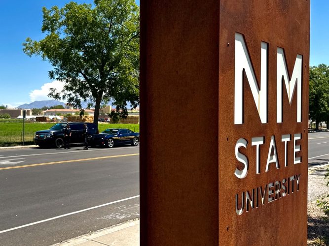 New Mexico State University sign near University Avenue, with Las Cruces Police and New Mexico State Police vehicles conducting a traffic safety operation in the background.