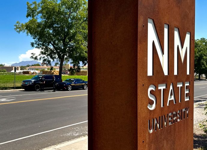New Mexico State University sign near University Avenue, with Las Cruces Police and New Mexico State Police vehicles conducting a traffic safety operation in the background.
